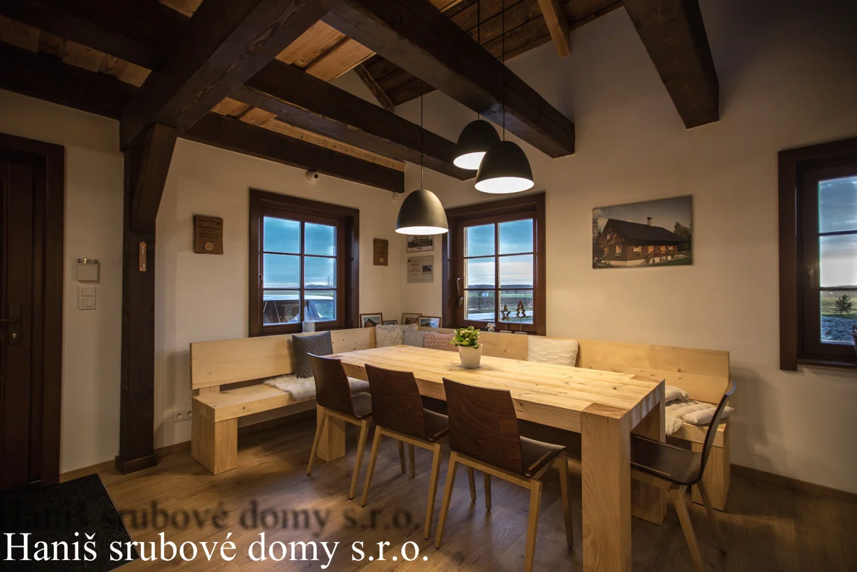 Jídelní stůl s lavicemi pod trámovým stropem, na zdi fotografie srubu